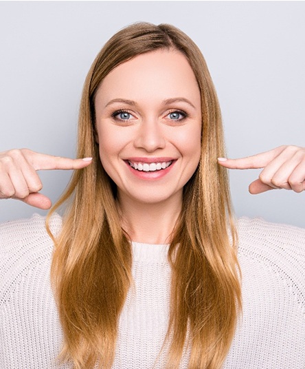 A young woman wearing a cream-colored sweater and pointing to her smile after completing treatment with Invisalign in Alhambra