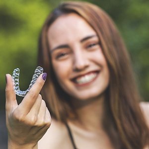 A young female smiling in the background and holding her Invisalign aligner between her thumb and forefinger